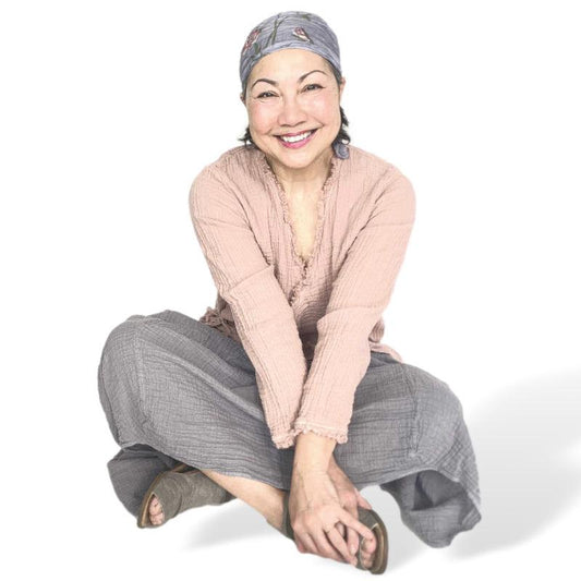 Smiling woman wearing comfortable cotton gauze pink long sleeve top and long grey skirt. She is sitting on the floor cross legged.