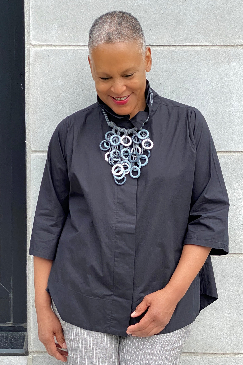 Older woman wearing a stylish outfit standing outside on a city street. She is wearing a black blouse with a silver and grey multi loop necklace.