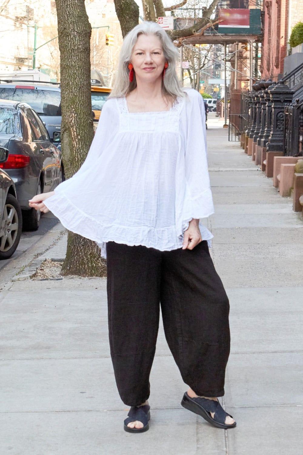 Woman with grey long hair wearing a curved shaped white cotton blouse with detaila and ruffule hemline. Worn with a black linen full cut pants. 