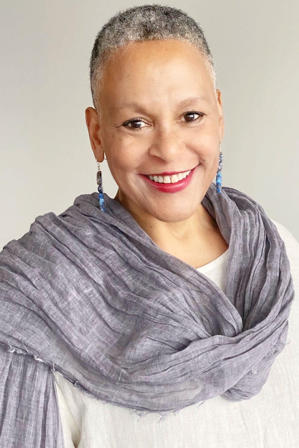 Lavender Grey cotton scarf wrapped around the neck of a smiling older woman with cropped grey hair. She is wearing blue earrings and a soft white cotton tee.