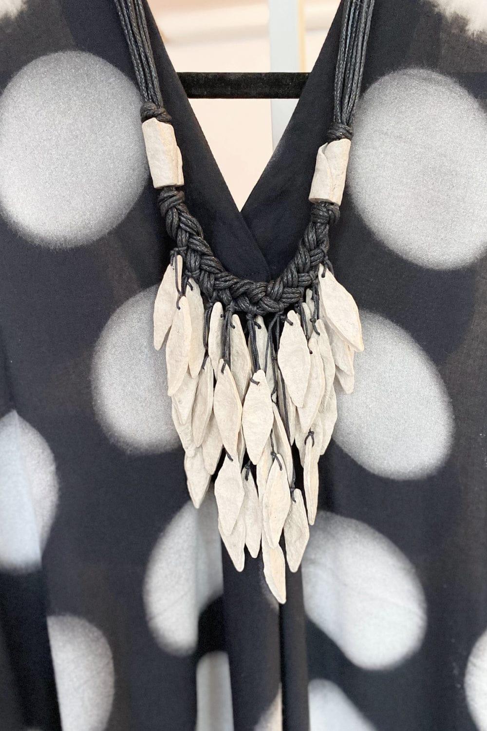 Silver colored sliver shapes cascading from woven a black woven cord. Two silver colored beads rest on each side of cord and bead and loop back closure. Worn with a black and off white polka dot kaftan
