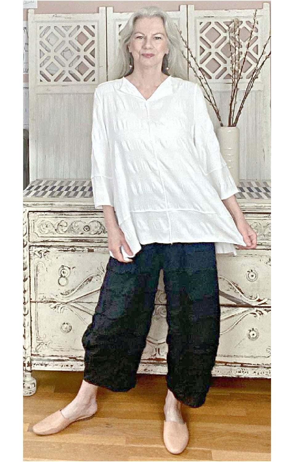 Aline shaped top with a split neckline and three quarter sleeves. Stylew with textured black full cut lantern pants. Worn on a older woman with long grey hair.